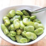 Bowl of cooked bright green fava (broad) beans with salt and pepper in a white bowl with a spoon.