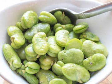 Bowl of cooked bright green fava (broad) beans with salt and pepper in a white bowl with a spoon.
