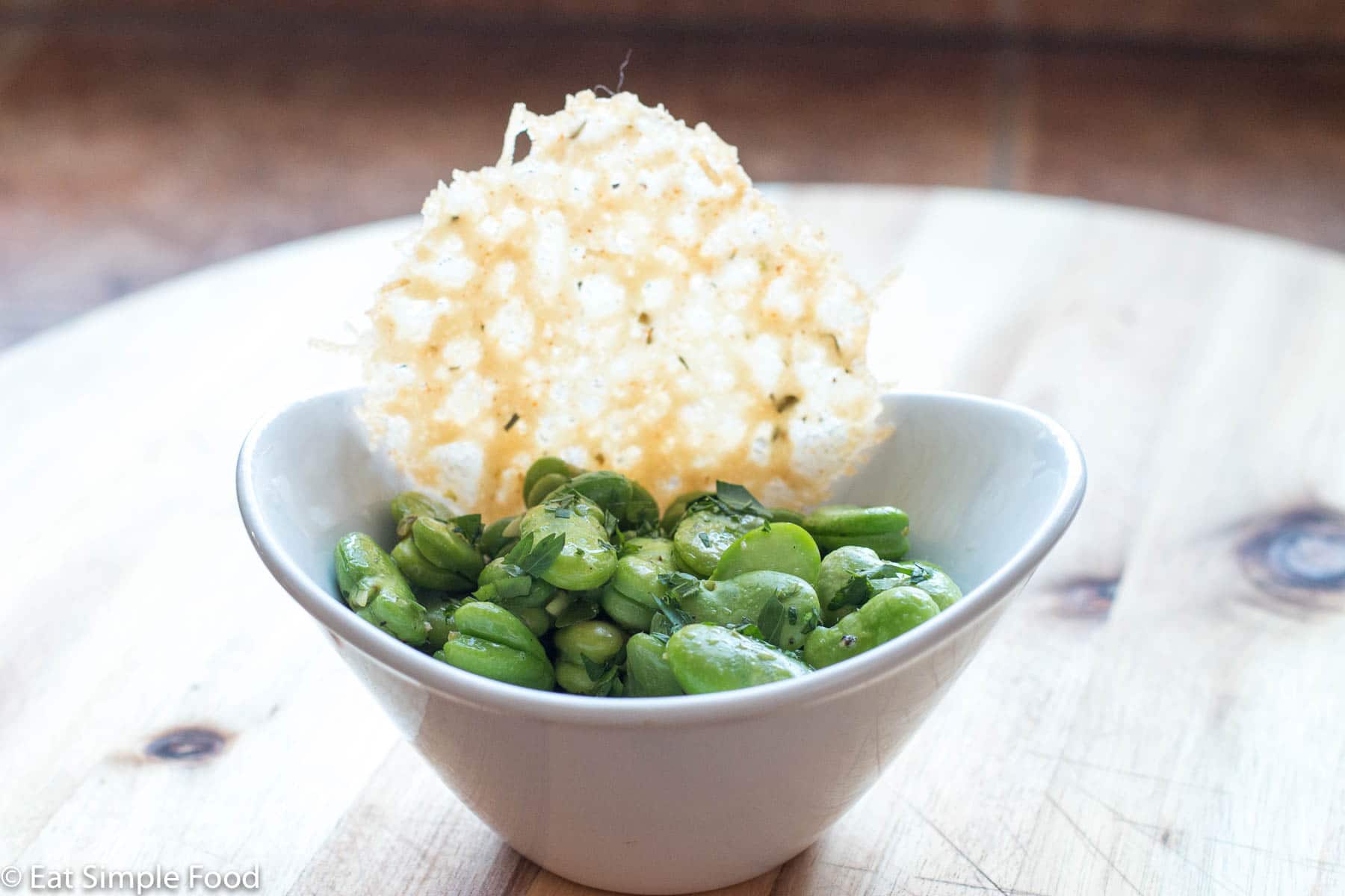 Bowl of cooked bright green fava (broad) beans with salt and pepper in a white bowl with a spoon. topped with mint. garnished with a parmesan crisp.