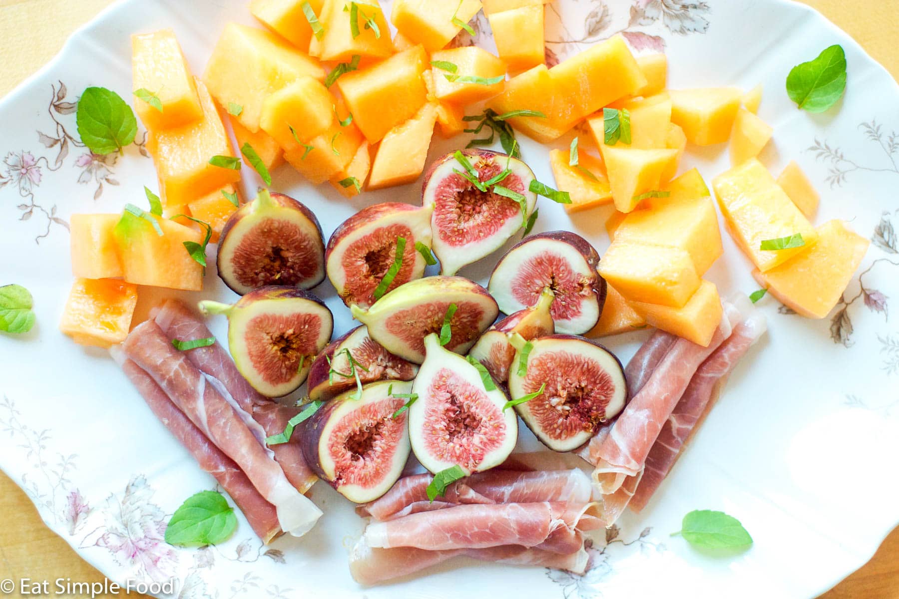 Halved figs, rolled prosciutto, chunks of cantaloupe on a platter with small mint leaves.