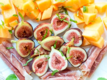 Halved figs, rolled prosciutto, chunks of cantaloupe on a platter with small mint leaves.
