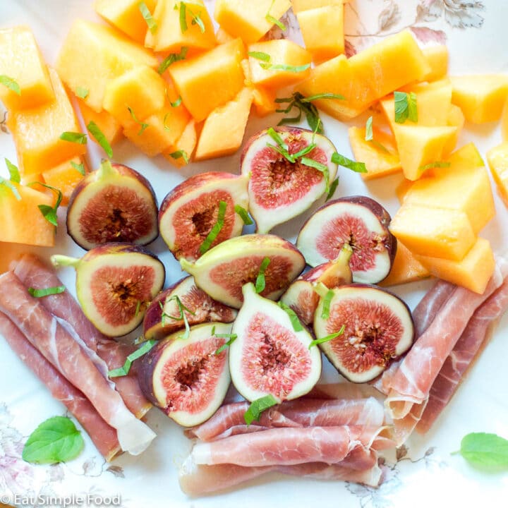 Halved figs, rolled prosciutto, chunks of cantaloupe on a platter with small mint leaves.