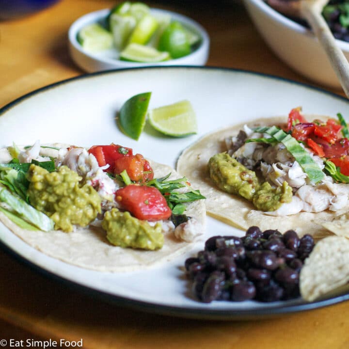2 soft shell tacos on a plate filled with fresh red tomatoes, guacamole, cabbage slaw with 2 wedges of lime and black beans on the side.