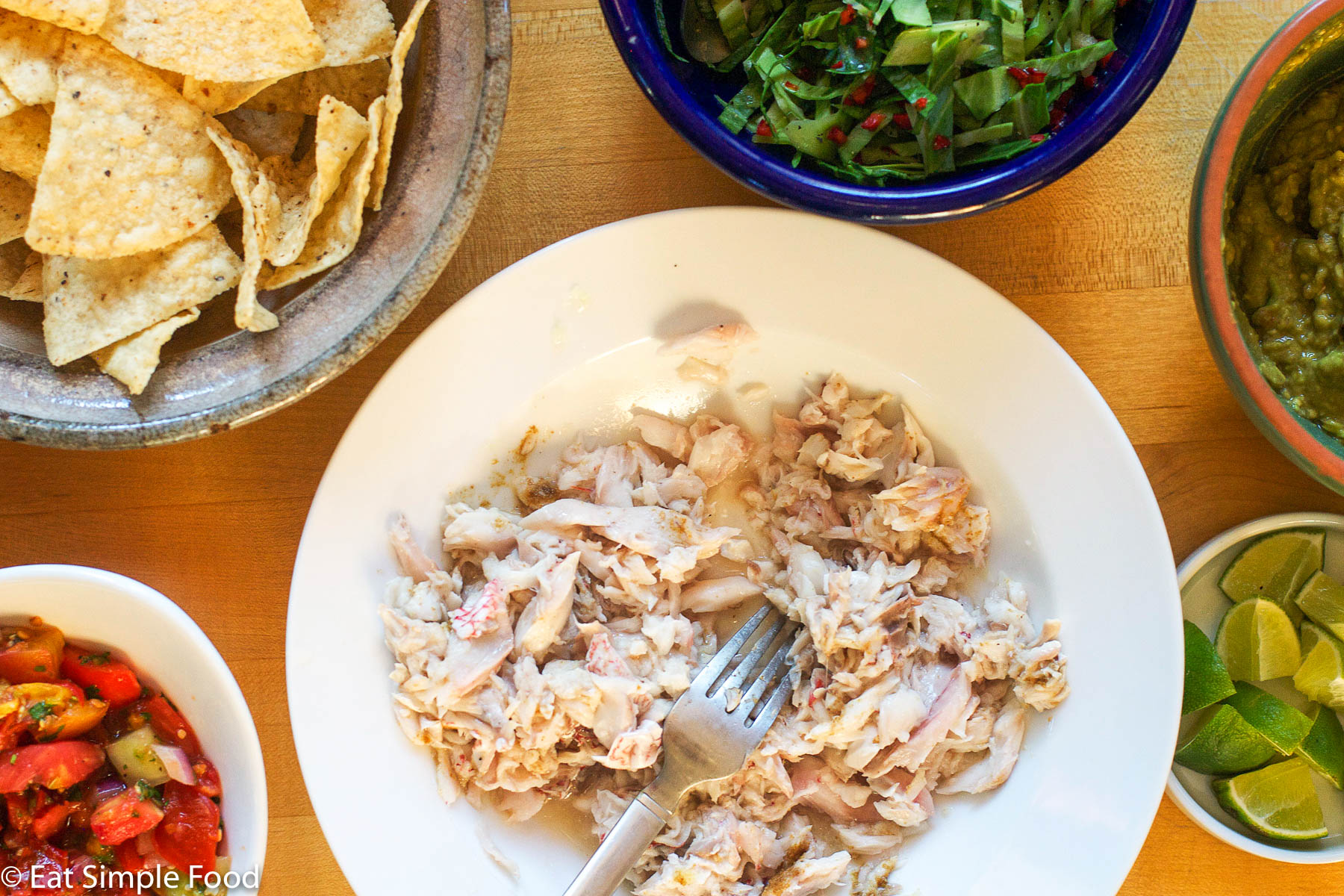 Ingredients for fish tacos on plates or bowls: cooked fish, lime wedges, cabbage slaw, guacamole, tortilla chips, tomato salsa