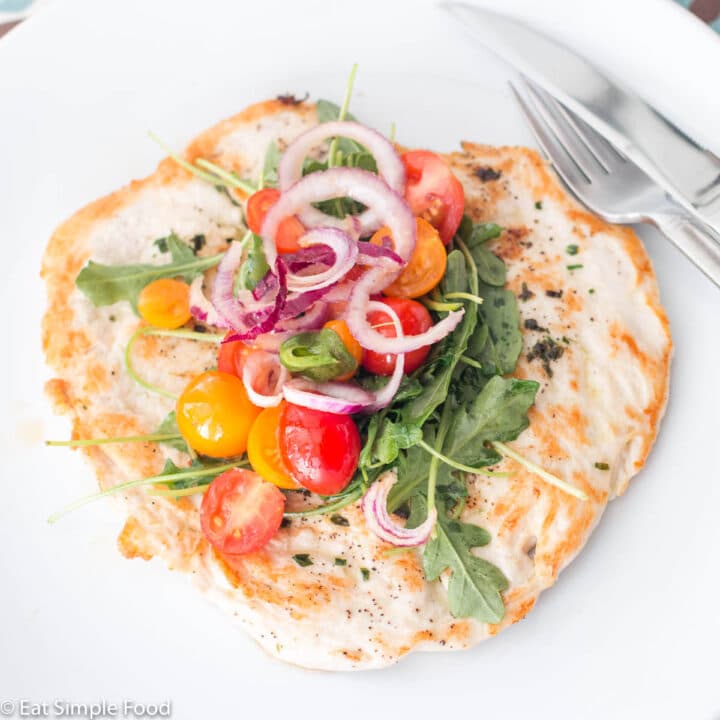 Flattened and seared chicken breast topped with Arugula salad w/ cherry tomatoes & onions on a white plate. Side view.