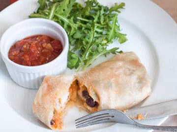 Burrito on a plate cut open to expose black beans, eggs, and cheese. Side of tomato salsa and arugula. Fork and knife on plate.