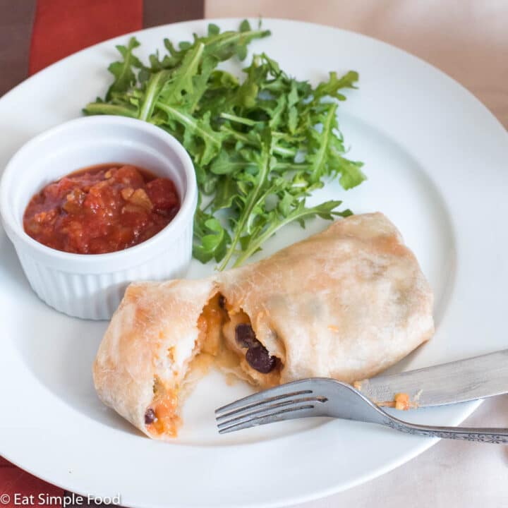Burrito on a plate cut open to expose black beans, eggs, and cheese. Side of tomato salsa and arugula. Fork and knife on plate.