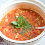 White bowl of ½ blended and ½ chunky cucumber and tomato gazpacho soup with basil leaf garnish