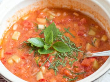 White bowl of ½ blended and ½ chunky cucumber and tomato gazpacho soup with basil leaf garnish