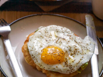 Fried egg on top of a piece of fried bread with a little avocado on a plate with knife and fork.