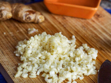 Minced garlic on a wood cutting board with a small orange square container. Knife and ginger knob on side.
