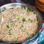 Top view. Stainless steel pan filled with thin noodles in a light soy sauce with garlic, chopped peanuts, and sliced green onions.