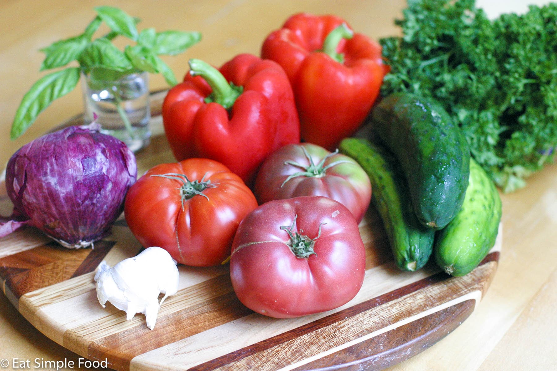 Gazpacho ingredients on a round wood cutting board: cucumbers, red peppers, garlic, tomatoes, red onion, parsley, and basil.