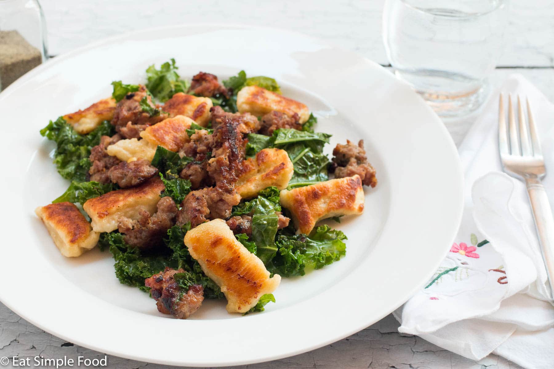 ¾ of a plate filled with browned gnocchi, sautéed kale, and ground sausage. On a white plate with a white napkin with small colorful flowers.