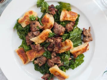 ¾ of a plate filled with browned gnocchi, sautéed kale, and ground sausage. On a white plate with a white napkin with small colorful flowers. Top view.
