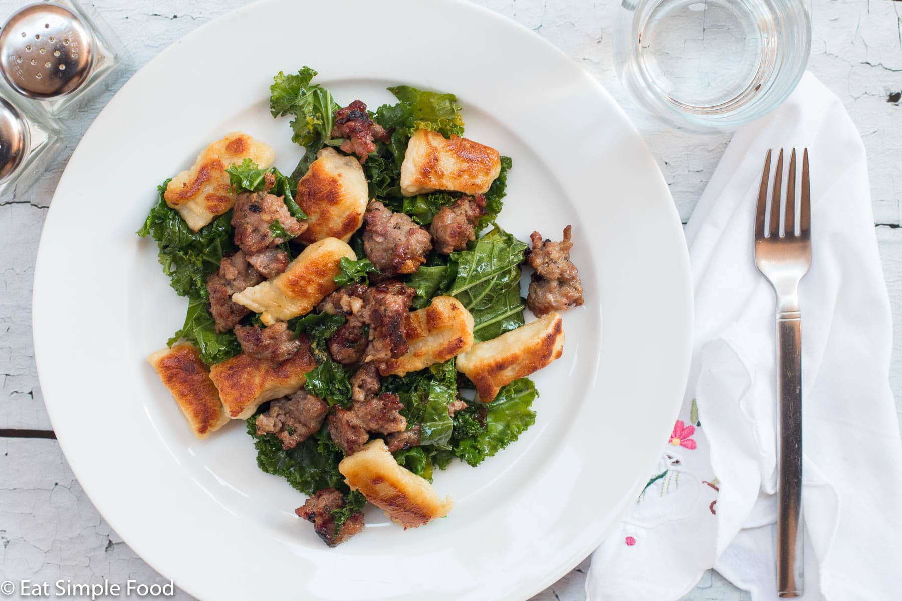 ¾ of a plate filled with browned gnocchi, sautéed kale, and ground sausage. On a white plate with a white napkin with small colorful flowers. Top view.