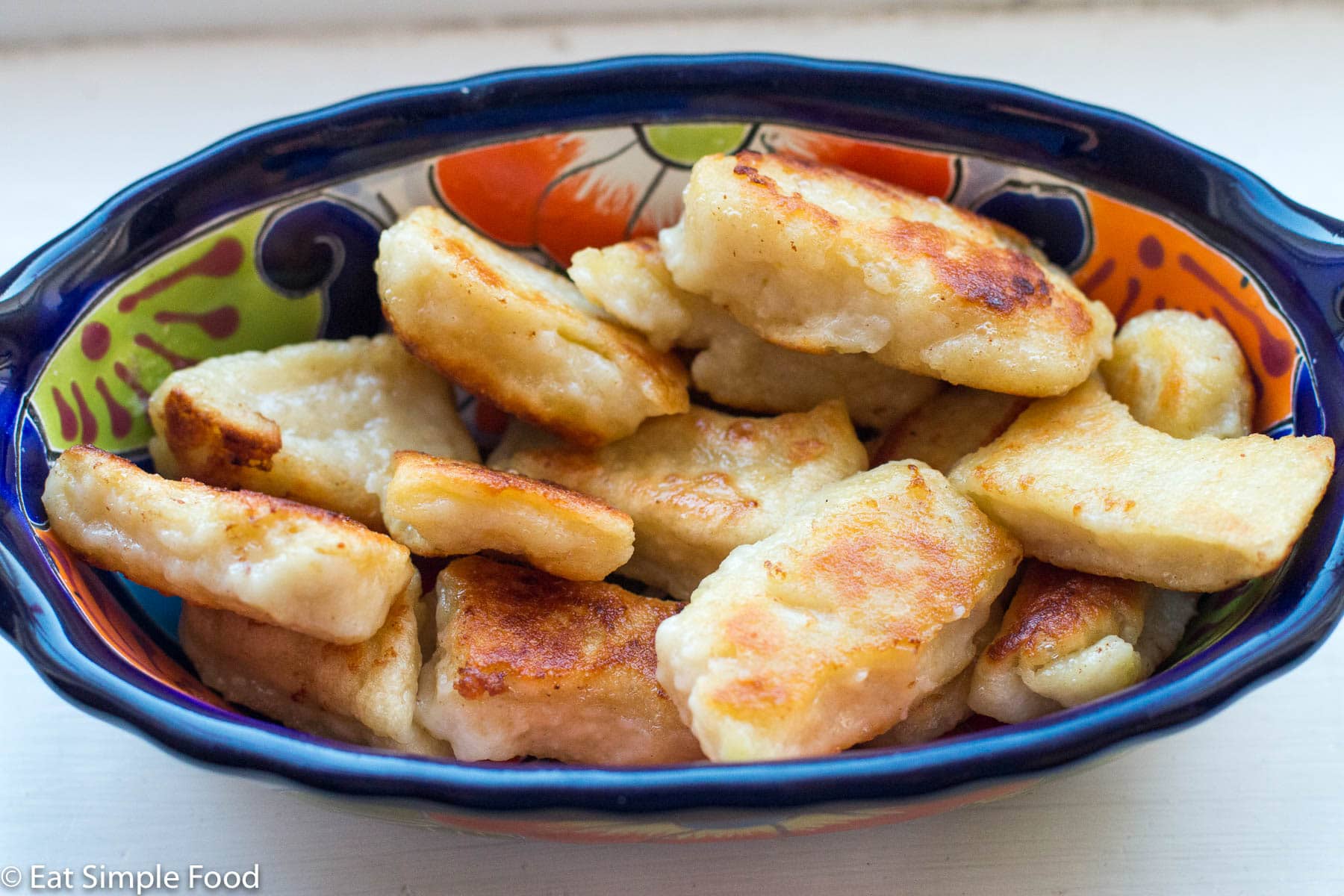 Blue oval bowl of golden brown pan fried gnocchi dumplings. On a white table.