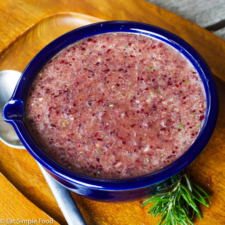 Blue Bowl of Purple Grape Sauce on a wood plate with a sprig of rosemary and a silver spoon.