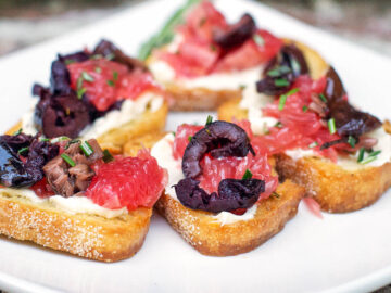 5 pieces of toasted baguette slices with mascarpone, grapefruit slices, sliced kalamata olives, and fresh rosemary on top.