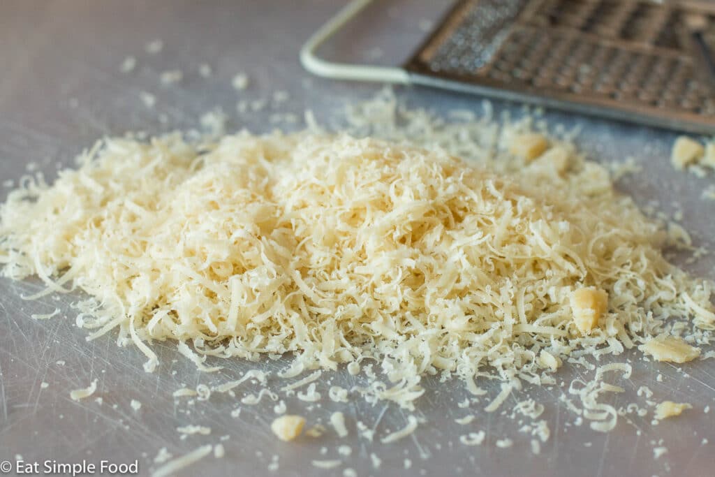 grated cheese on a see-through cutting board with a hand held cheese grater in the background.