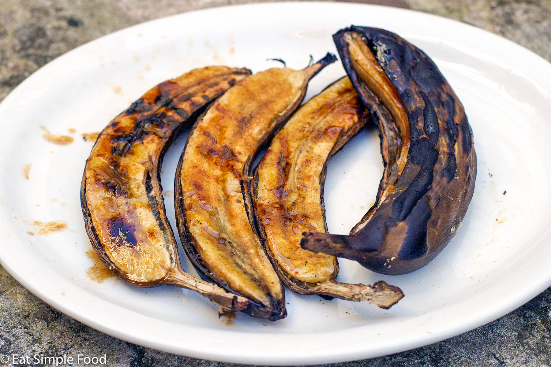 3 Grilled Banana Halves and One Whole, still in skin & grilled. Set on a white plate and has a honey butter glaze.