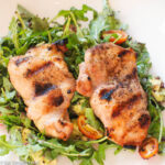 2 grilled chicken thighs over a bed of arugula salad w/ halved cherry tomatoes and diced avocado. On a white plate.