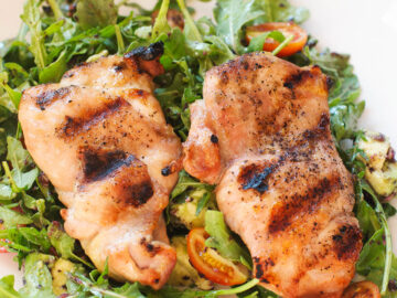 2 grilled chicken thighs over a bed of arugula salad w/ halved cherry tomatoes and diced avocado. On a white plate.