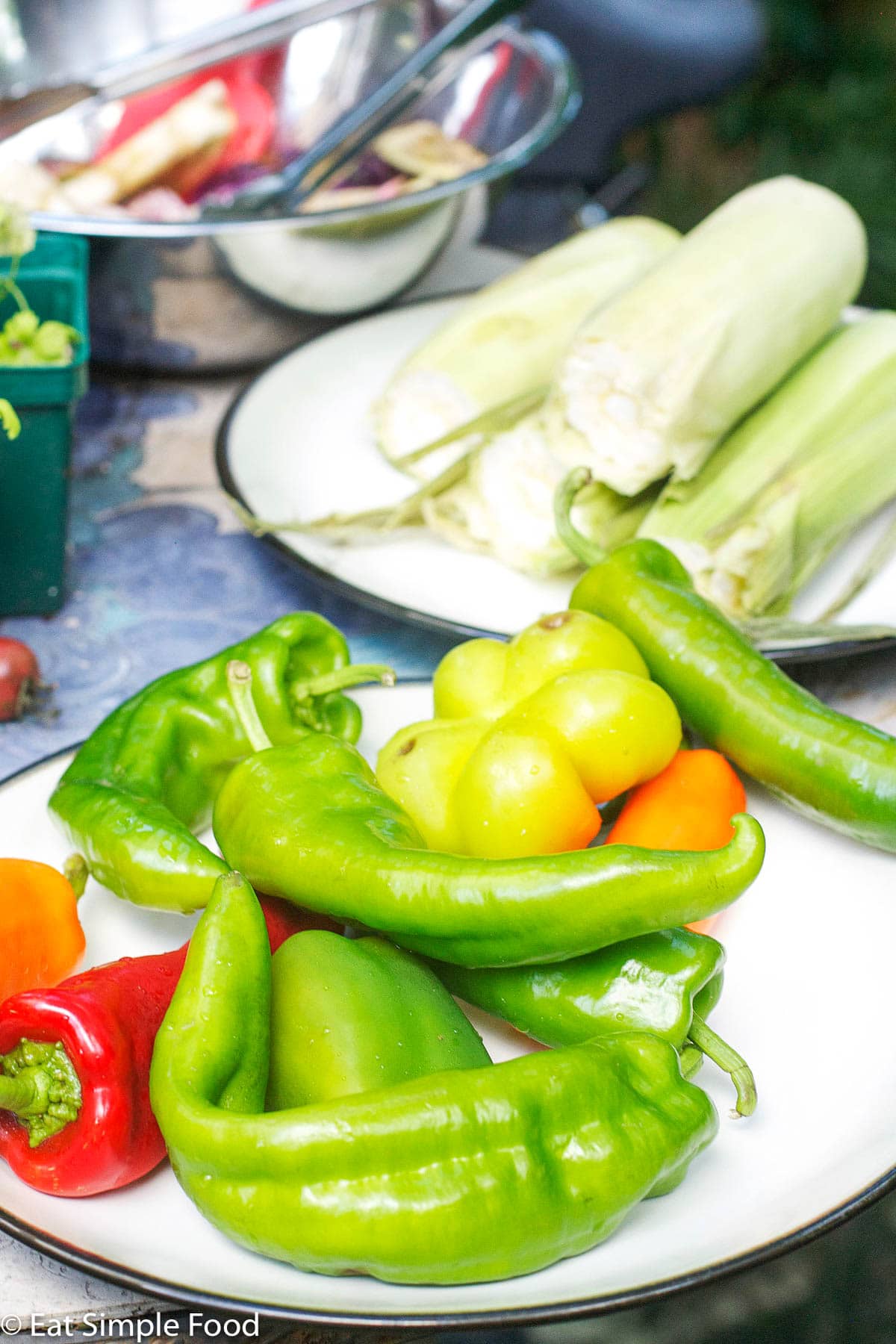 Red, Orange, and green poplano peppers on a white plate with a plate of corn with husk in the background.