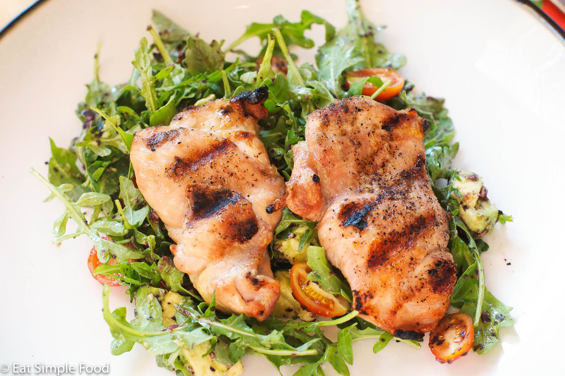 2 grilled chicken thighs over a bed of arugula salad w/ halved cherry tomatoes and diced avocado. On a white plate.