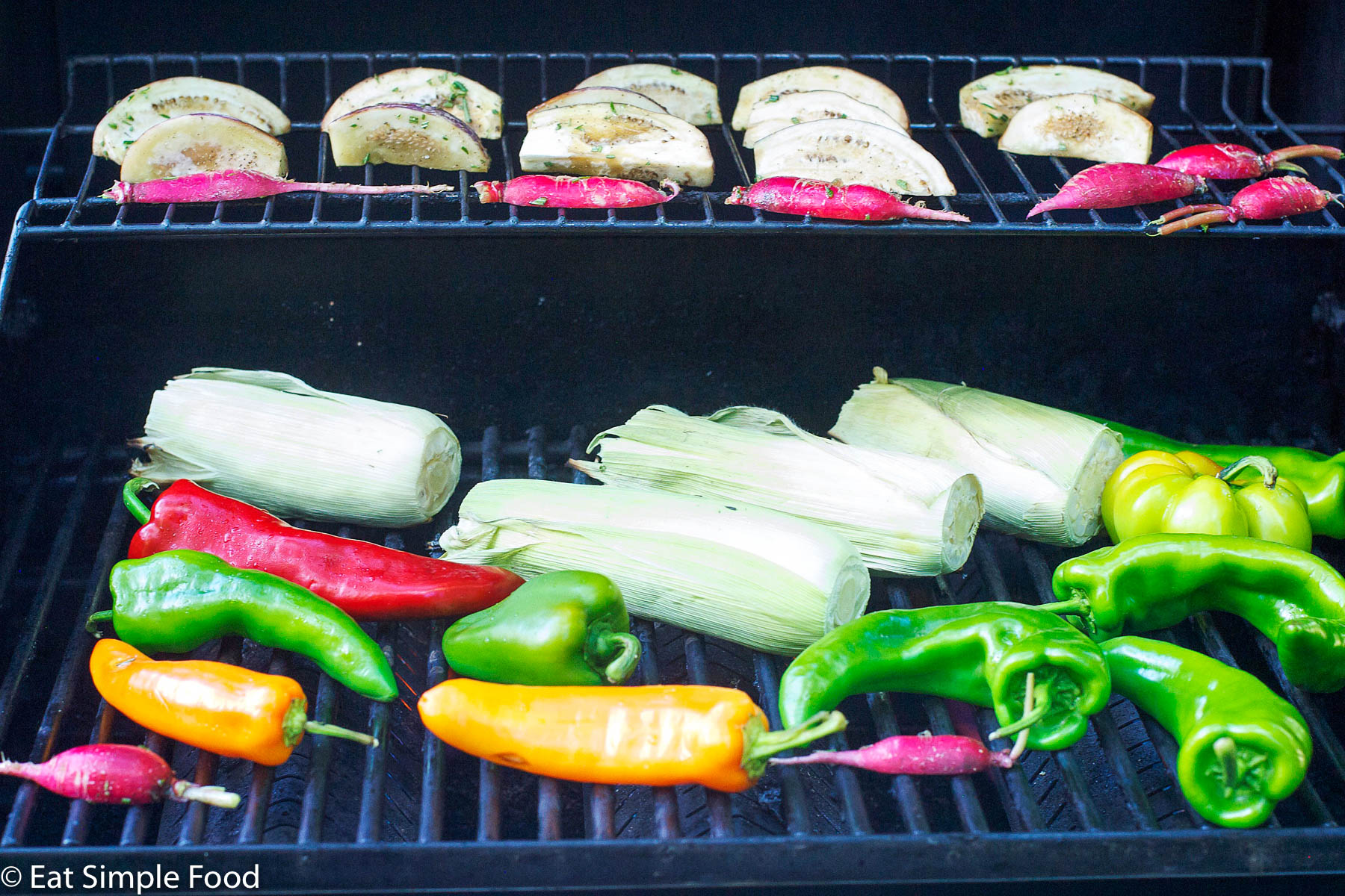 Corn on the cob in husk, whole poblano peppers (green, red, and orange), quartered eggplant and small red peppers on two levels of a grill