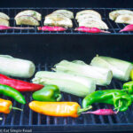 Corn on the cob in husk, whole poblano peppers (green, red, and orange), quartered eggplant and small red peppers on two levels of a grill