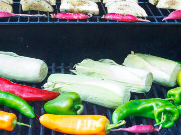 Corn on the cob in husk, whole poblano peppers (green, red, and orange), quartered eggplant and small red peppers on two levels of a grill