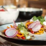2 Ground beef tacos in flour tortillas filled with tomatoes, guacamole, goat cheese, sliced radishes, peppers and cilantro. Bowl of salsa in background.