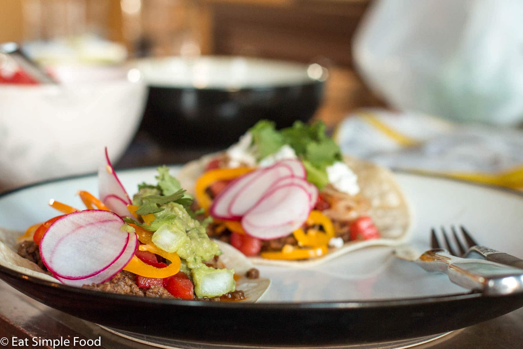 2 Ground beef tacos in flour tortillas filled with tomatoes, guacamole, goat cheese, sliced radishes, peppers and cilantro. Two bowls in background.