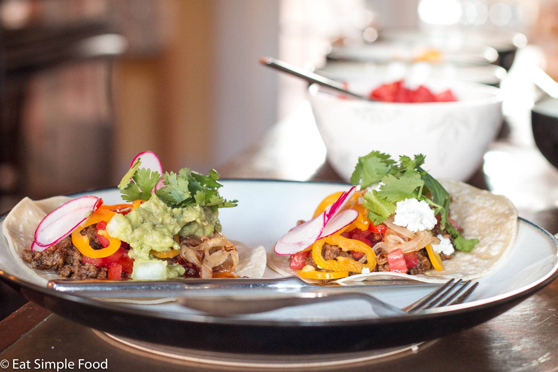 2 Ground beef tacos in flour tortillas filled with tomatoes, guacamole, goat cheese, sliced radishes, peppers and cilantro. Bowl of salsa in background.