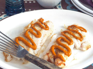 Side view of pan seared white fish with red romesco sauce drizzled on the top. Fish spatula on plate.