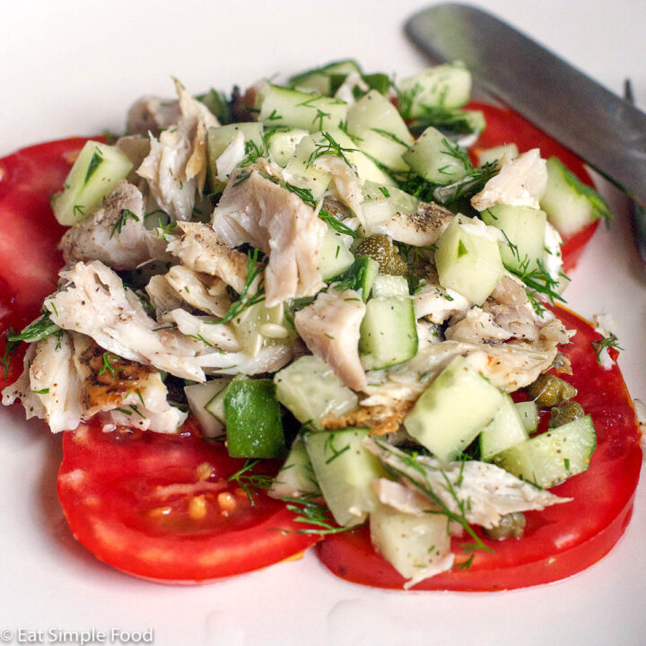 White plate with a layer of sliced tomatoes at the bottom. On top is diced grouper and cucumbers with fresh dill and capers.