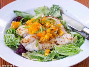 Seared piece of white fish and a bed of romaine lettuce with chunked yellow tomato and sliced basil salsa. White plate with knife and fork on the side.