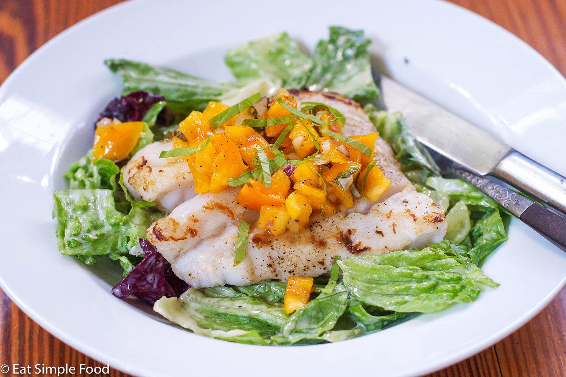 Seared piece of white fish and a bed of romaine lettuce with chunked yellow tomato and sliced basil salsa. White plate with knife and fork on the side.