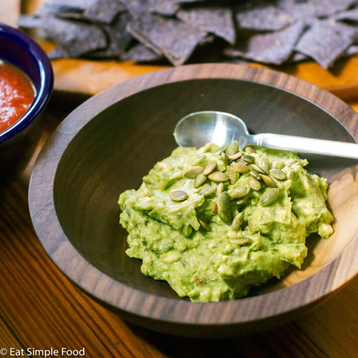 Guacamole in a wood bow with toasted pepita garnish with a spoon in bowl. Blue tortilla chips and bowl of salsa in background.