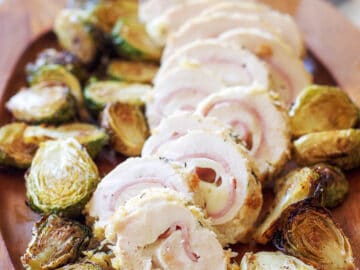 Chicken, Ham, & Cheese Roulades (Rolls) sliced into rounds on a wood plate with roasted brussels sprouts.