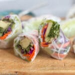2 spring rolls cut in half (4 halves) filled with avocado, grated carrots and red cabbage, rice noodles, green lettuce on a wood cutting board.