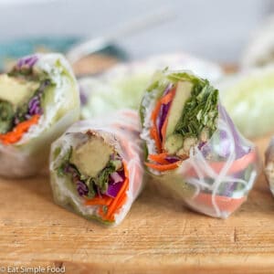 2 spring rolls cut in half (4 halves) filled with avocado, grated carrots and red cabbage, rice noodles, green lettuce on a wood cutting board.