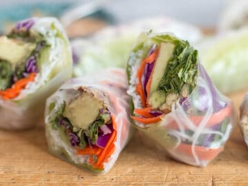 2 spring rolls cut in half (4 halves) filled with avocado, grated carrots and red cabbage, rice noodles, green lettuce on a wood cutting board.