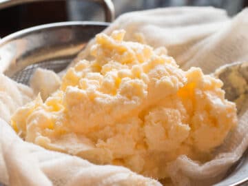Homemade yellow Butter in an open cheesecloth over a sieve with a butter scraper.