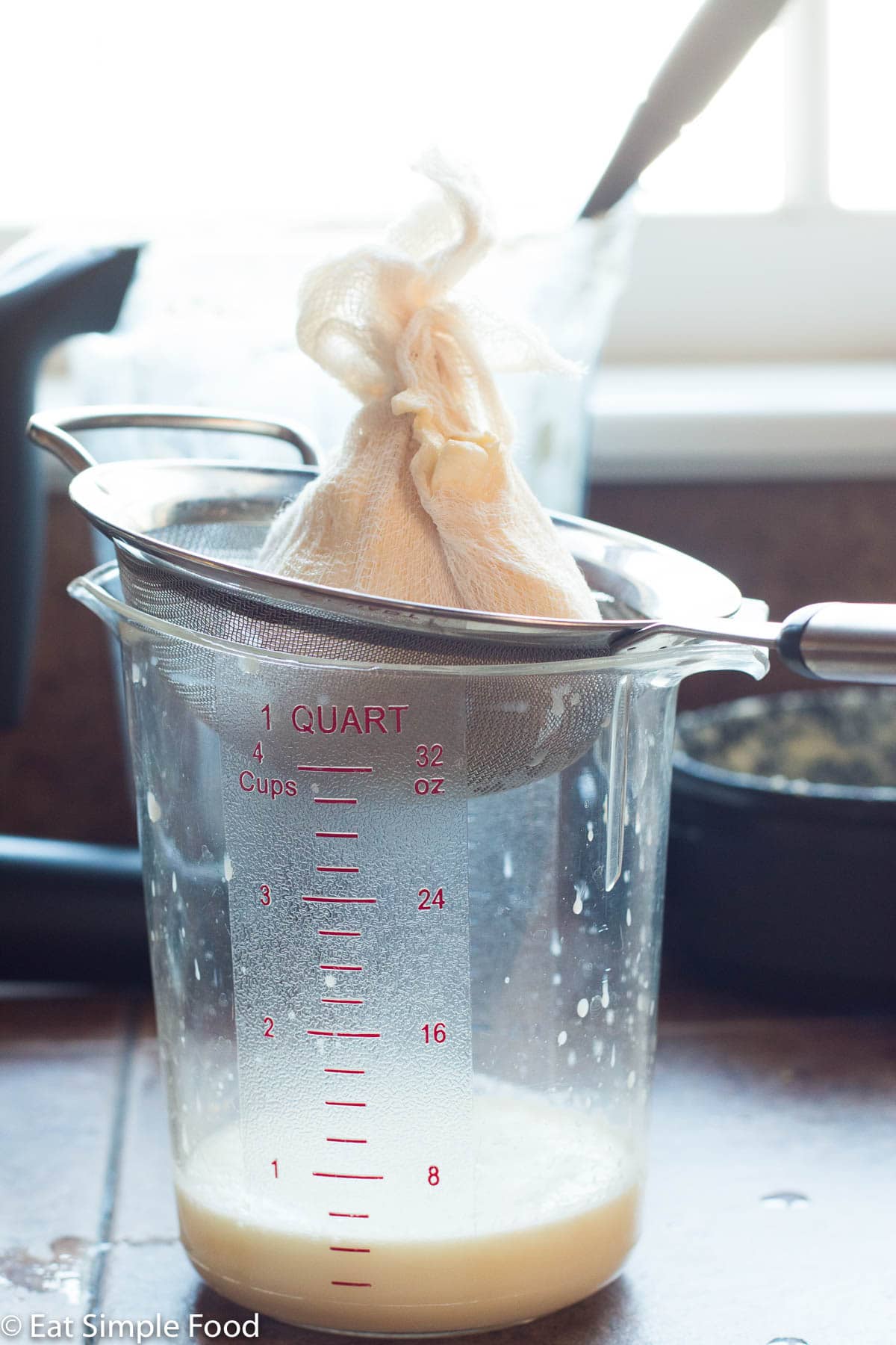 A sieve on top of a 4 cup plastic clear measuring cup. The sieve has homemade butter wrapped in cheesecloth being drained.