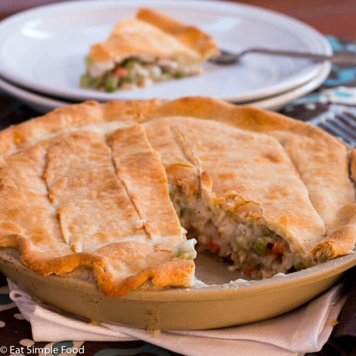 Whole golden brown chicken pot pie with one slice missing. Pie slice on white plate in background.