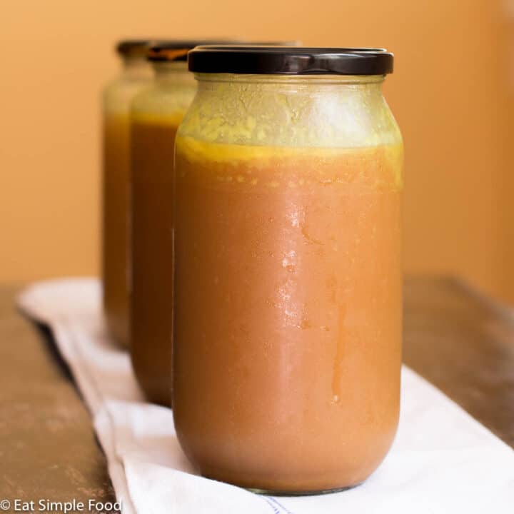 3 Mason Jars Lined up vertically with rich brown chicken stock in them on a white kitchen towel.