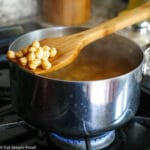 Stainless steel pot of dry garbanzo beans cooking over an open flame with a wooden spoon sticking out.
