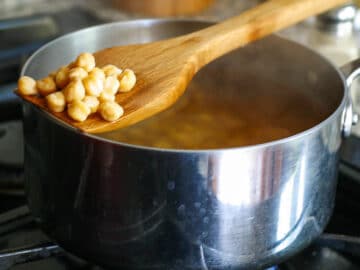 Stainless steel pot of dry garbanzo beans cooking over an open flame with a wooden spoon sticking out.